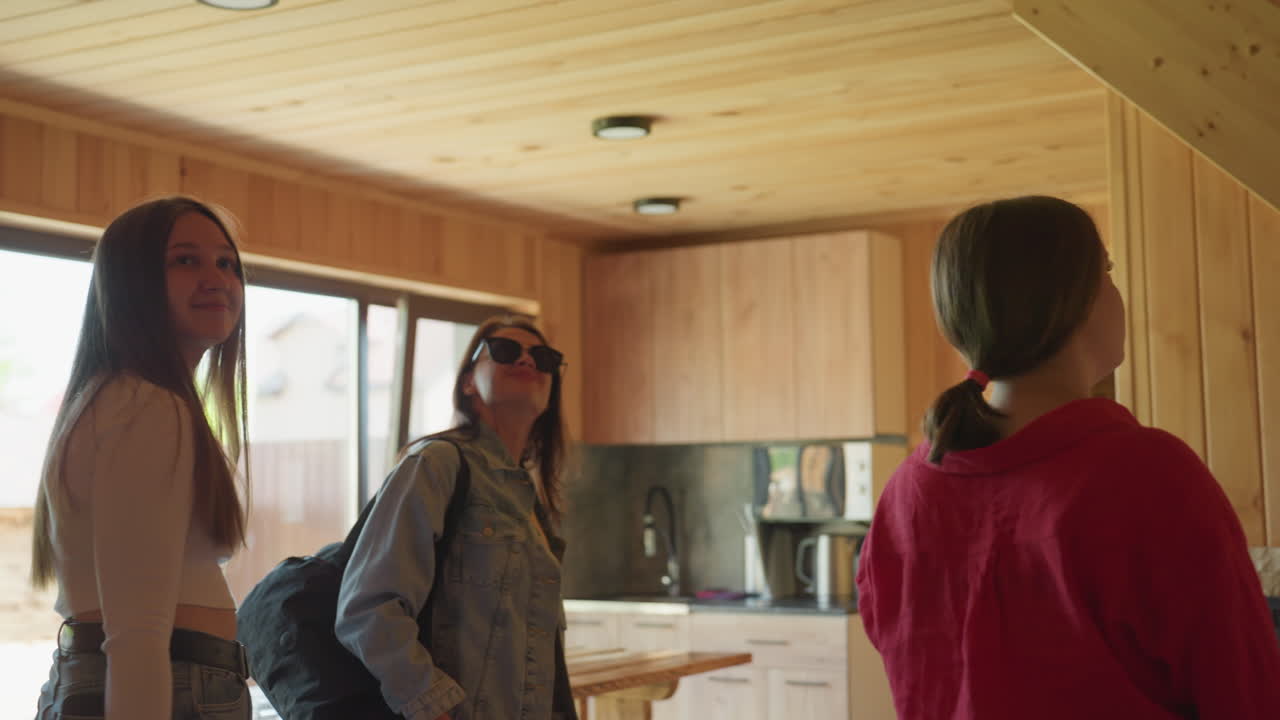 Dimly lit view of wooden apartment interior with mounted television as group of ladies enter and observe space, capturing quiet exploration moment filled with anticipation and curiosity