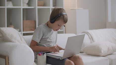 school boy is drawing at home and talking by internet with friend using laptop and headphones with microphone