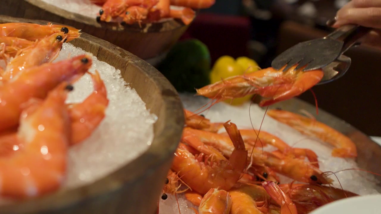A hand uses metal tongs to select chilled prawns from a wooden bowl on ice