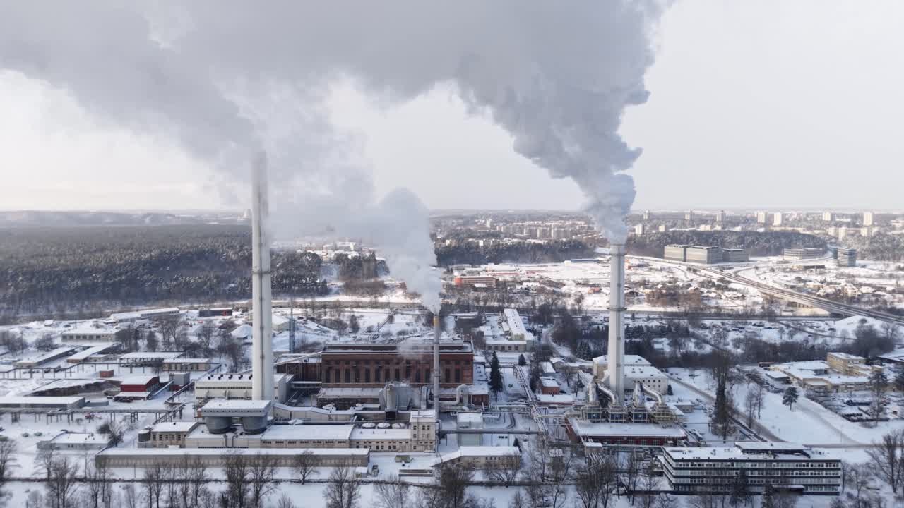 Drone view of Vilnius cogeneration facility with tall smokestacks releasing white vapor above snowy industrial district and forest hills, Lithuania