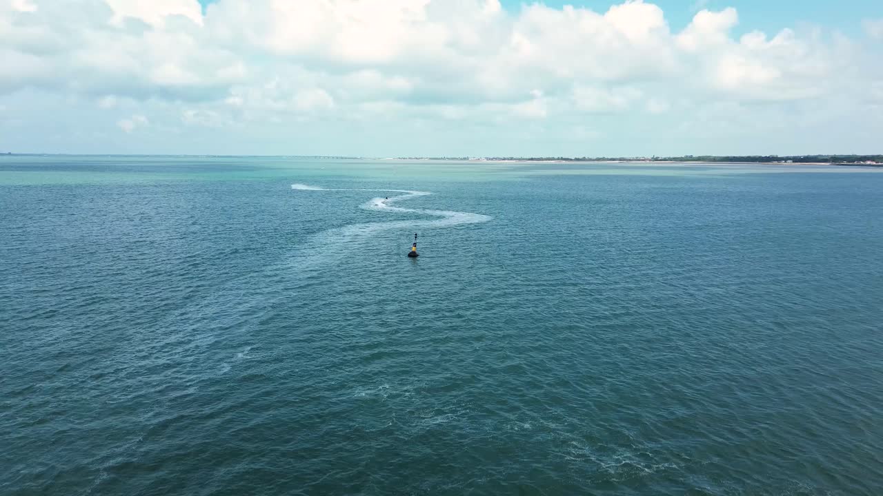 Sweeping aerial view of jetskiers speeding across the open ocean, contrasted against the rugged coastline and bright blue horizon of Charente-Maritime