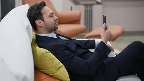 smiling middle-aged businessman lying on the sofa using and looking at his phone, man in suit watching social media, scrolling, browsing the internet sitting in living room couch
