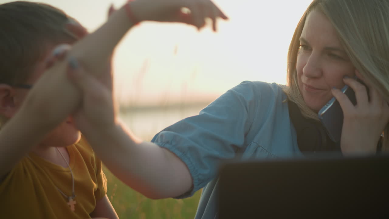 A woman in a blue gown sits in a grassy field, holding a phone to her ear. She gently holds her son's hand,who is wearing a yellow outfit,preventing him from distracting her while she manages the call