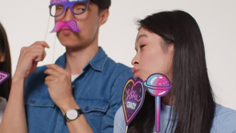 Side-View-Of-Group-Of-Young-Friends-In-Front-Of-White-Studio-Background-Having-Fun-Posing-For-Photo-Booth-Style-Portraits-With-Props-2