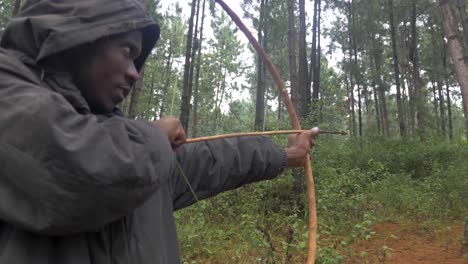 a slow motion orbit shot around a hooded african hunter shooting a traditional wooden bow and arrow in a forest