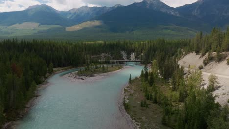 Absteigende-Drohnenaufnahme-Des-Türkisfarbenen-Gletscherwassers-Des-Kootenay-River-An-Einem-Sonnigen-Sommertag-Mit-Einer-Brücke-Im-Blick-Und-Forststraßen-Mit-Bergen-Im-Hintergrund