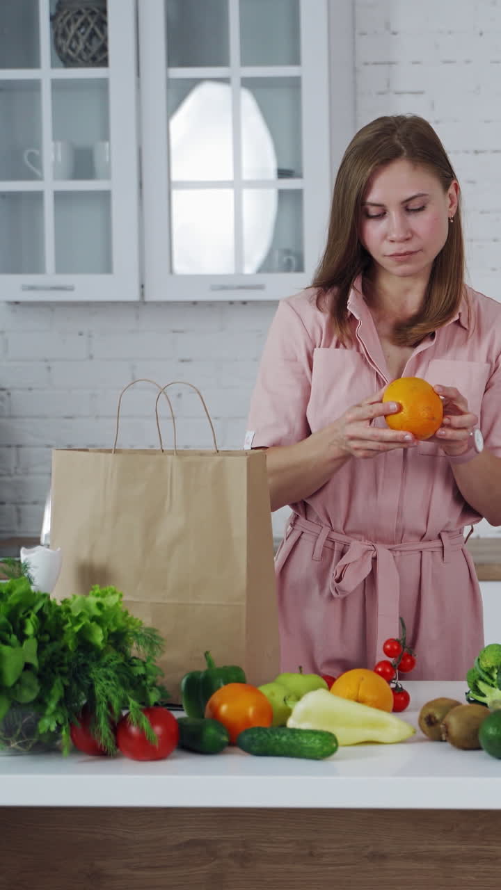 Pretty woman taking out fresh fruit from paper shopping bag at home. Young female holds an orange in the light kitchen. Healthy fruit and vegetables on the table. Vertical video