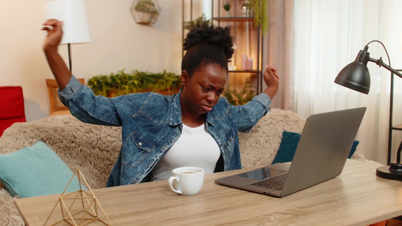 Giovane donna afroamericana beve stancamente il caffè prima di lavorare da remoto al computer portatile seduta a casa