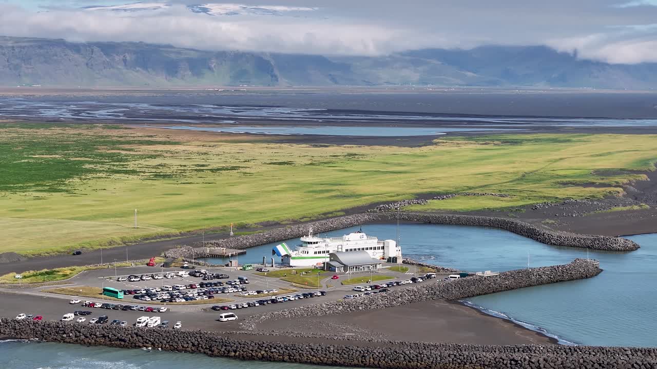 Electric car ferry arriving Iceland Island in summer. Aerial orbit shot. Parking cars on area of harbor. Green flat landscape and mountains in background. Volcanic island in Europe
