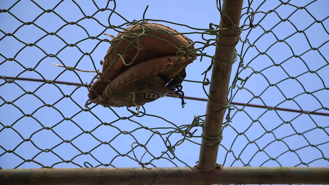 Luva de beisebol antiga presa em uma rede de proteção de corrente rasgada em um campo rural empoeirado na República Dominicana, simbolizando jogos esquecidos e tradições desportivas comunitárias desgastadas sob um céu azul claro