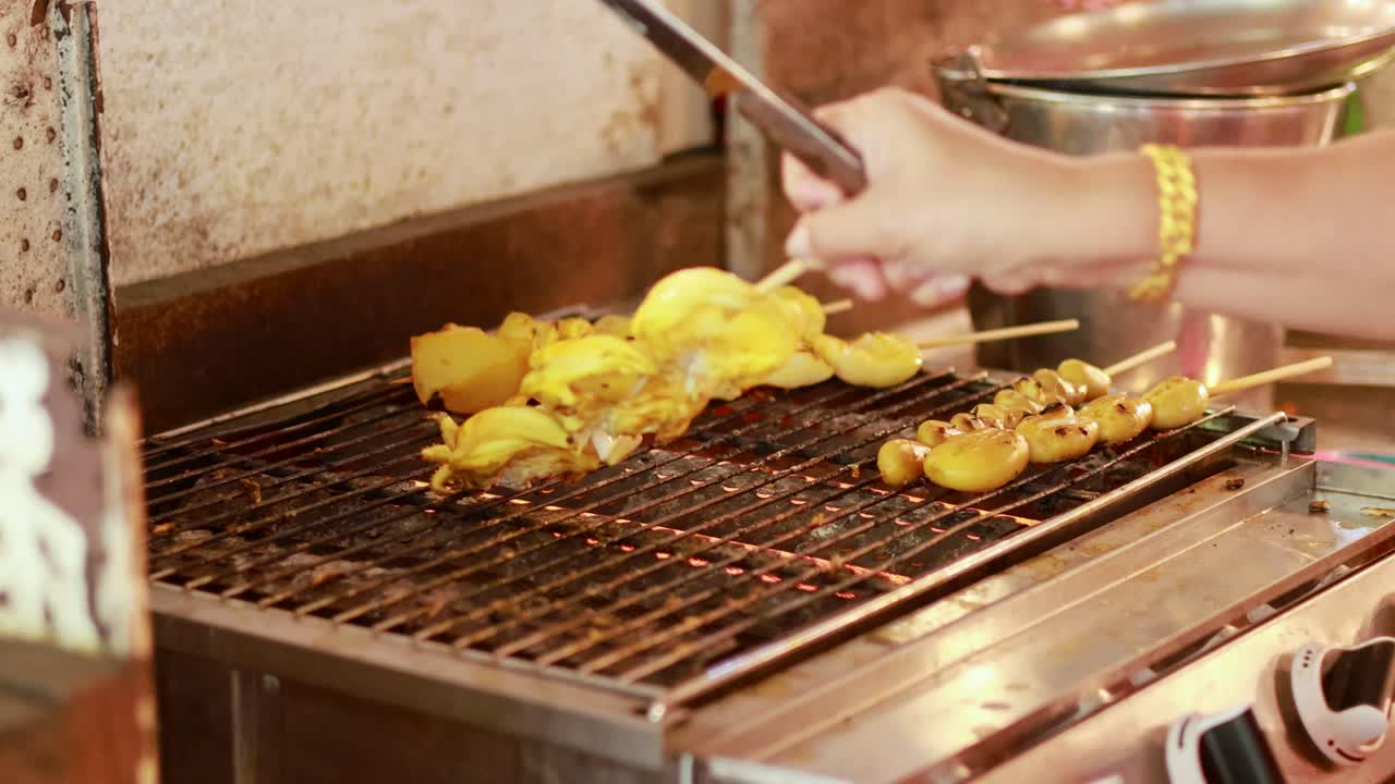 Hands grill skewered food on a metal barbecue at a bustling market. Warm lighting enhances the vibrant, lively atmosphere