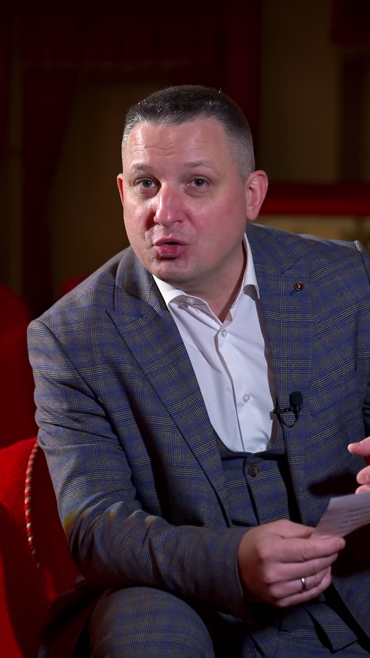 Performer announces future theater season. Actor is sitting in empty theater on red velvet chair with sheet of paper in hands. Vertical video