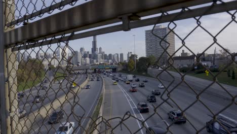 Toma-Desde-Una-Plataforma-Rodante-A-Través-De-Una-Valla-Metálica-Hasta-La-Concurrida-Autopista-Eisenhower-Y-El-Horizonte-De-Chicago