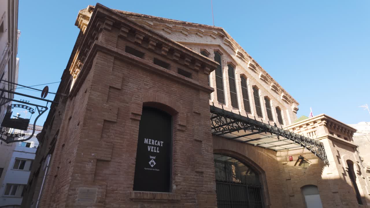 Low angle view of the Mercat Vell building in Sitges, featuring a brick facade, arched entrance, and ornate iron canopy