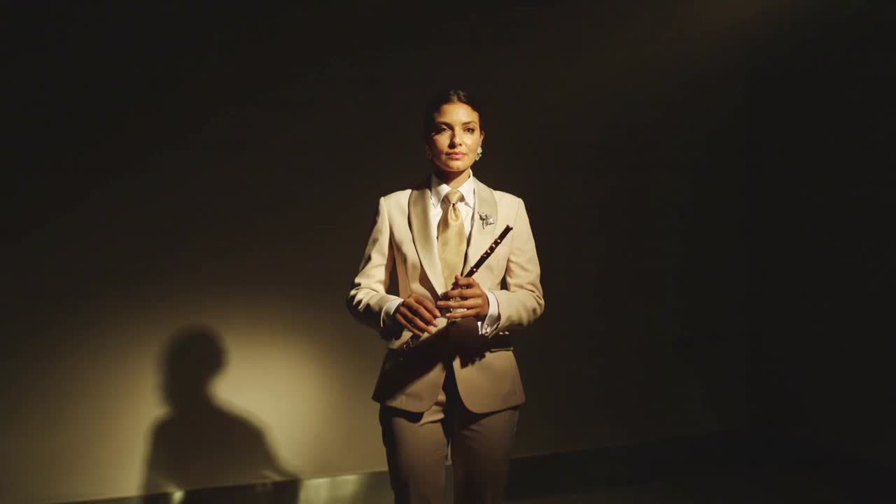Adjusting grip, female flutist in cream suit raising gold flute to lips, performing in dark studio
