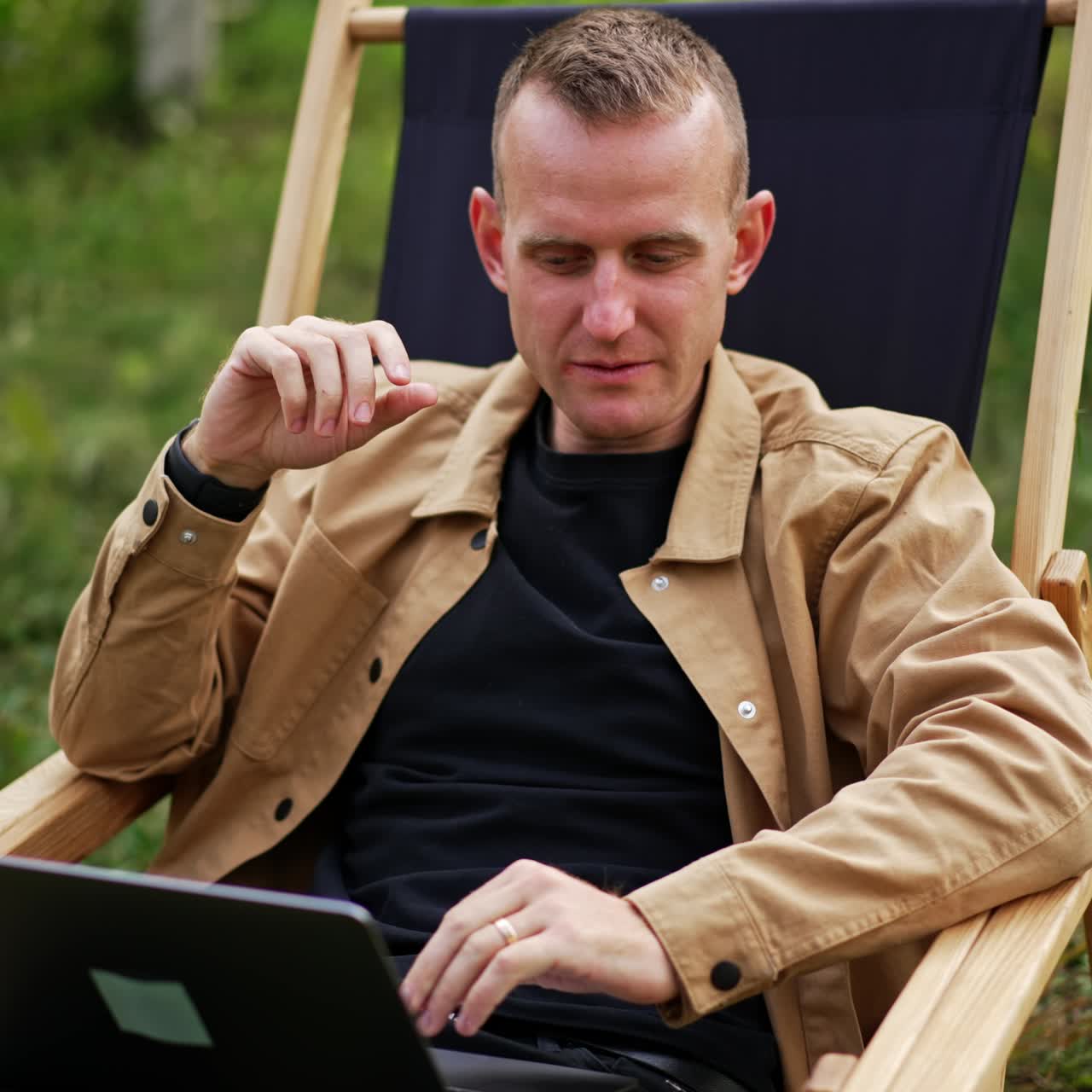 Positive relaxed man looks at the screen of his laptop. Male freelancer sitting comfortably in a garden chair working remotely from nature