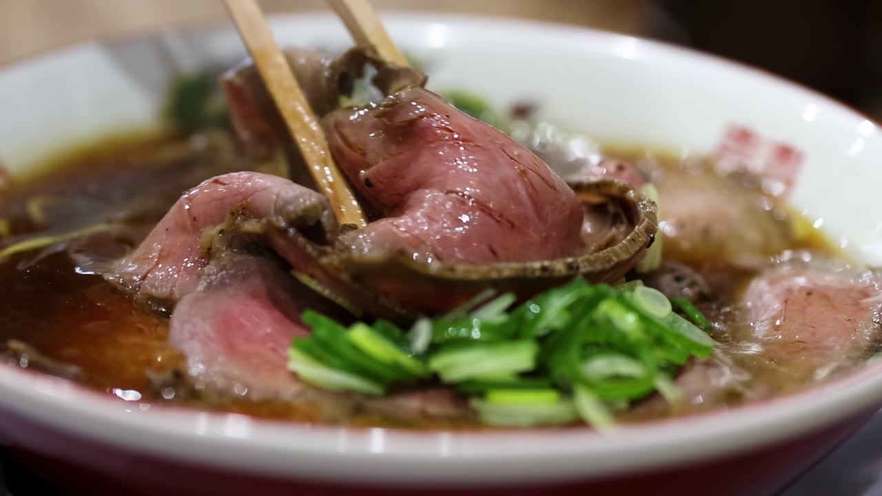 Close-up of noodles being lifted from a bowl with sliced meat and fresh green onions.