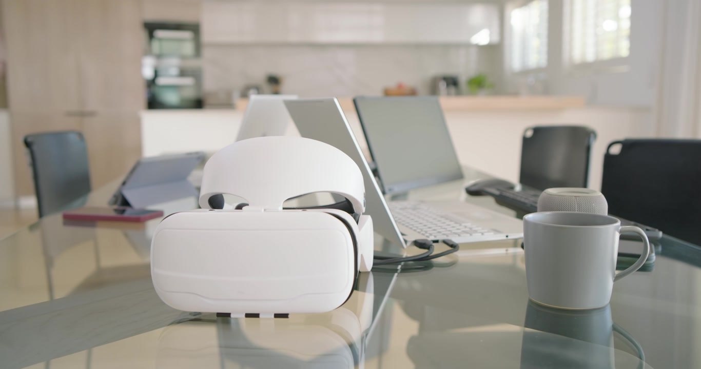 Camera is moving closer, causing VR headset to grow on table beside laptop, highlighting workspace