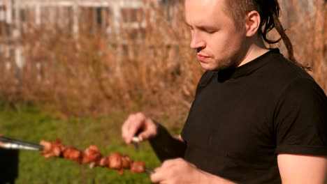 young man is eating meat shashlik barbecue on a skewer with knife in the backyard in the spring on a sunny day.