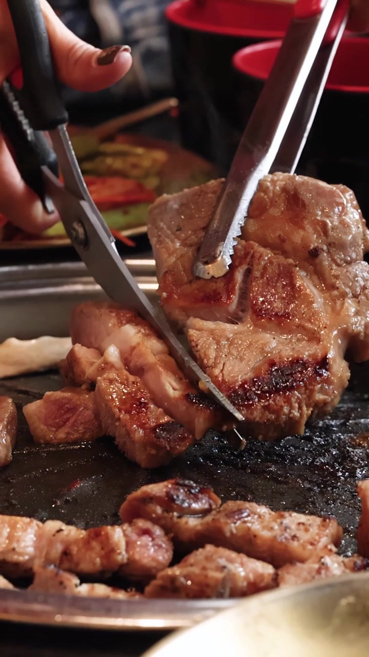 Tangan memotong daging babi panggang dengan gunting dan penjepit di atas panggangan barbekyu Korea di bawah pencahayaan hangat
