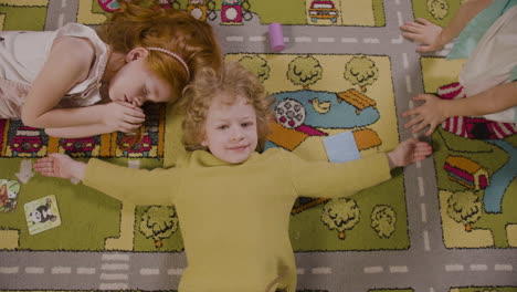 top view of a little boy lying on the carpet with open arms and looking at camera