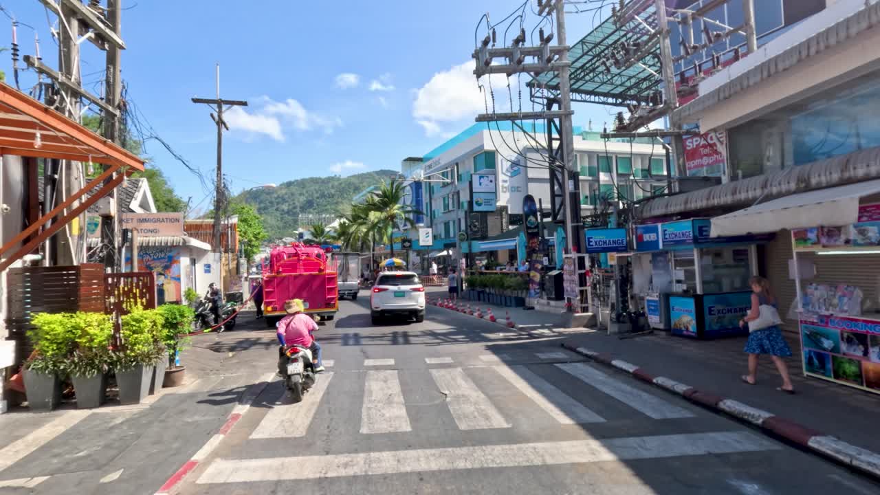 Daytime drive past shops, tuk-tuks, and scooters on busy Patong Beach road, clear weather