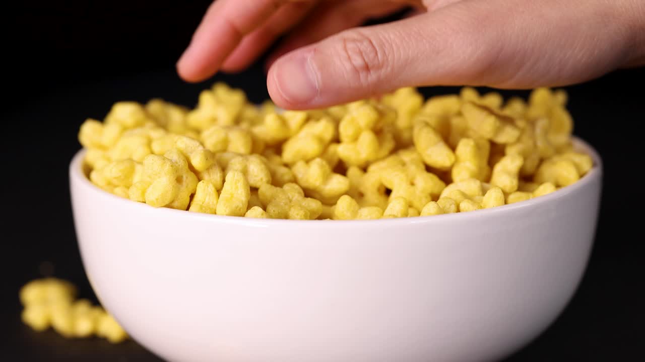 A hand explores and plays with yellow cereal pieces in a white bowl against a black background, highlighting texture and movement