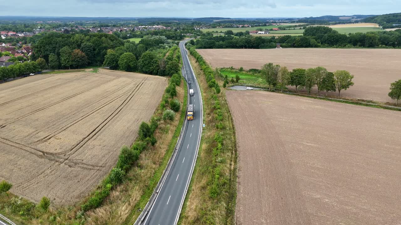 Truck on interstate highway between wheat fields in suburbia of city. Aerial flyover shot. Bridge leading to neighborhood of USA. Agricultural farm fields in rural district. Wide shot