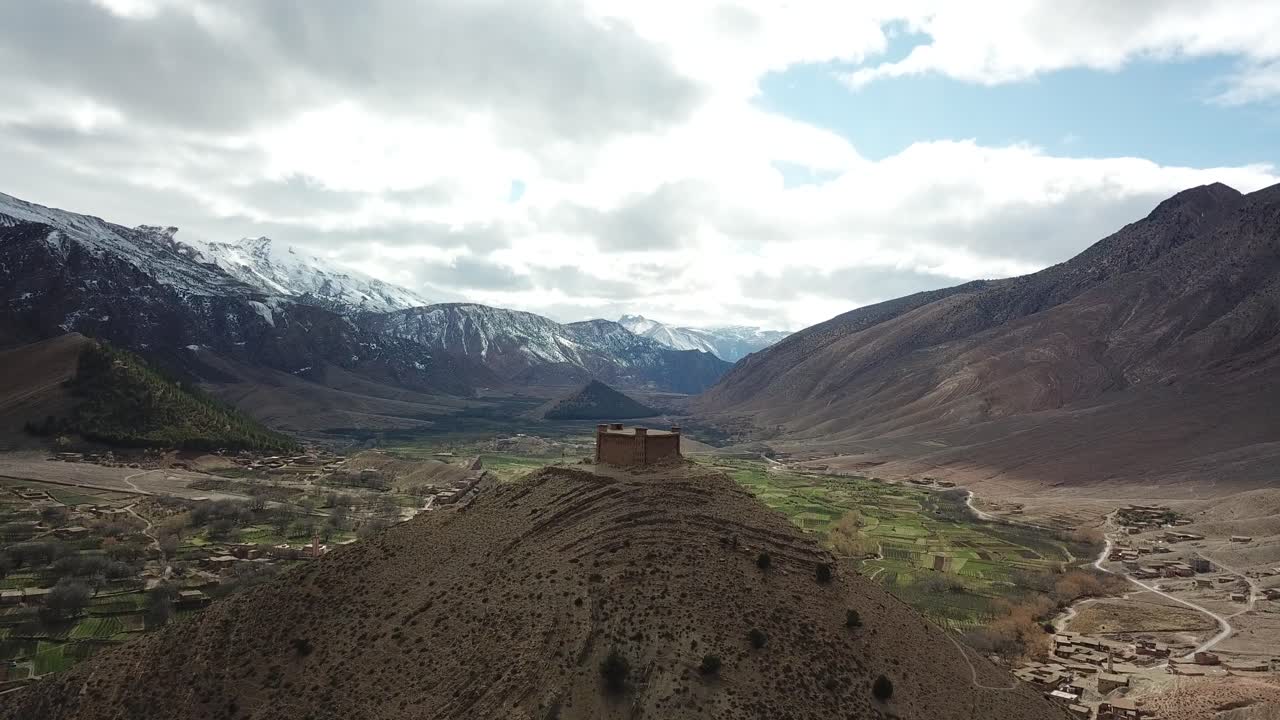An ancient kasbah crowns a hilltop in Morocco's Atlas Mountains, its red clay walls and defensive towers standing proud against dramatic peaks and endless mountain vistas