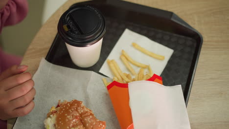 close-up of fries, a partially eaten burger, and a coffee cup on a black tray a brown wooden table, a woman s hand in a pink dress reaches for fries, takes a bite, and sips her coffee