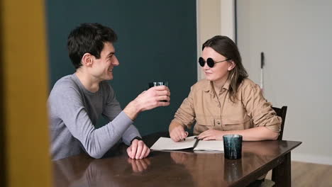 man and blind woman in sunglasses sitting at table at home talking