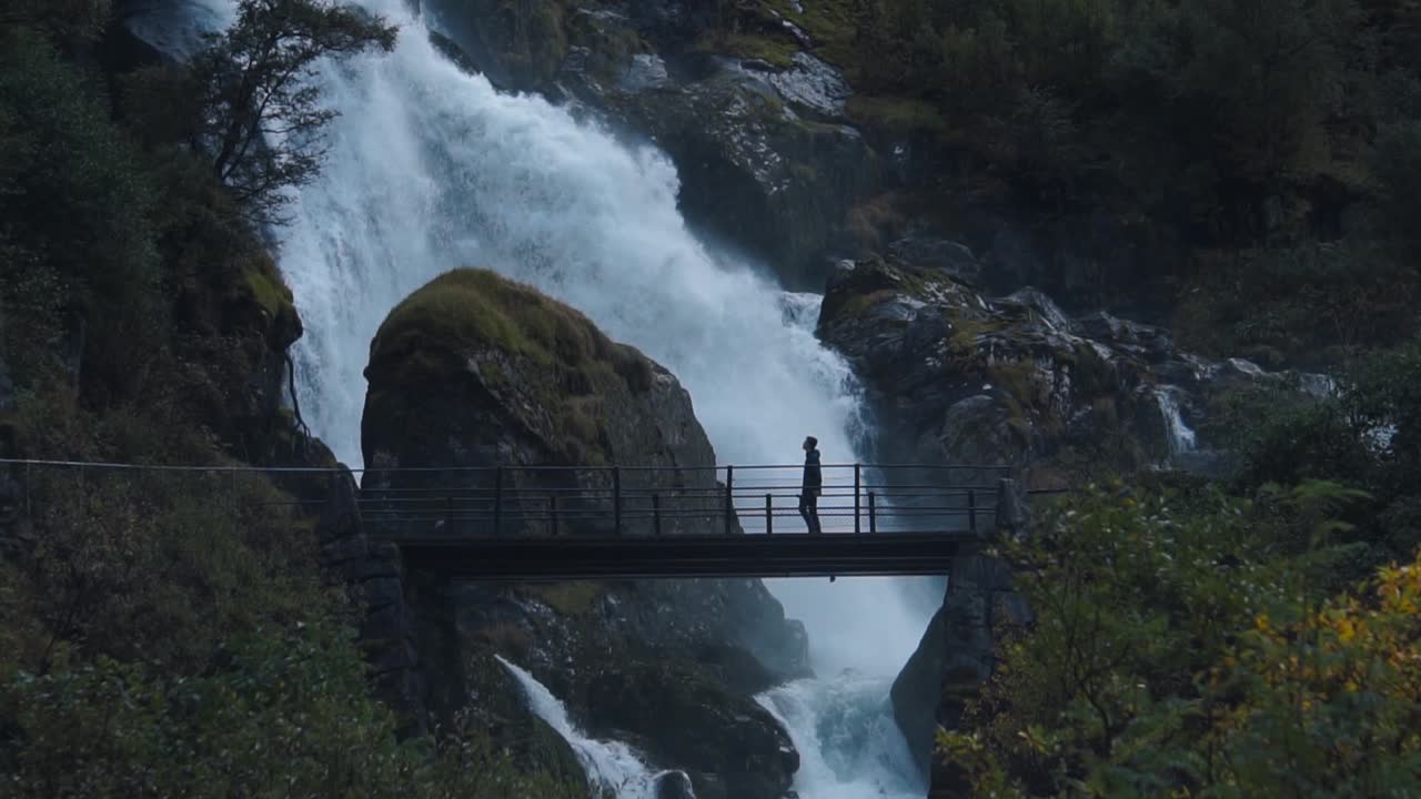 toma en cámara lenta de un hombre caminando frente a una cascada salvaje sobre un pequeño puente de madera, noruega briksdalen