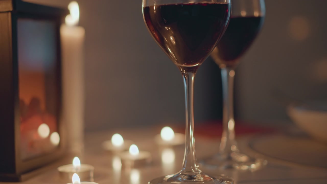 Vista em close-up de mesa de duas taças de vinho cheias de vinho tinto ao lado de velas tealight acesas e uma vela cônica em uma superfície brilhante; chamas suaves tremeluzem, fumaça leve flutua e reflexos quentes criam uma atmosfera acolhedora.