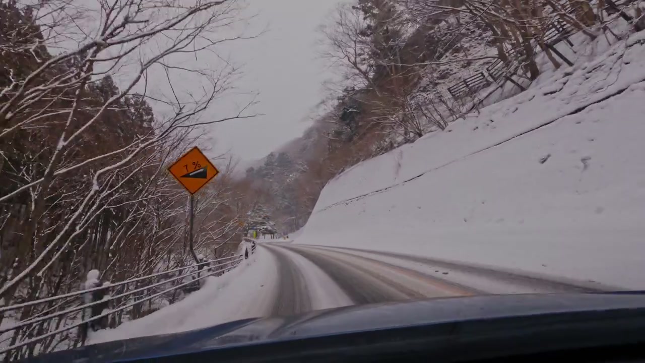 Dashcam-perspektiv navigerer på en smal, snesporet vej, der skærer gennem en dyb bjergdal. En betonviadukt strækker sig over kløften forude, indrammet af snedækkede skråninger og nøgne træer