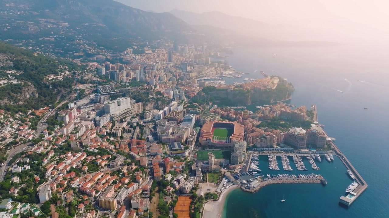 Le vedute aeree di Monaco catturano il paesaggio urbano con edifici, porto e costa. Si vedono barche nell'acqua mentre il sole tramonta sulle colline sullo sfondo.