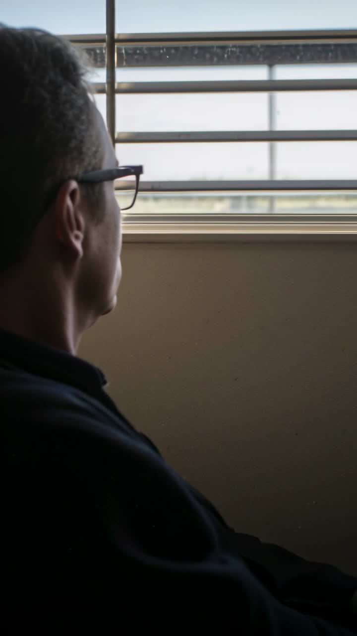 Vertical video: Man sitting office light shifting blinds gazing checking view fixing glasses