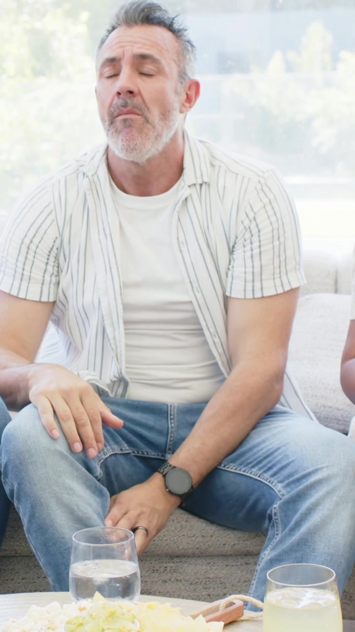 Vertical video: Reacting to comment, man in shirt reaching for pale drink composing himself on sofa