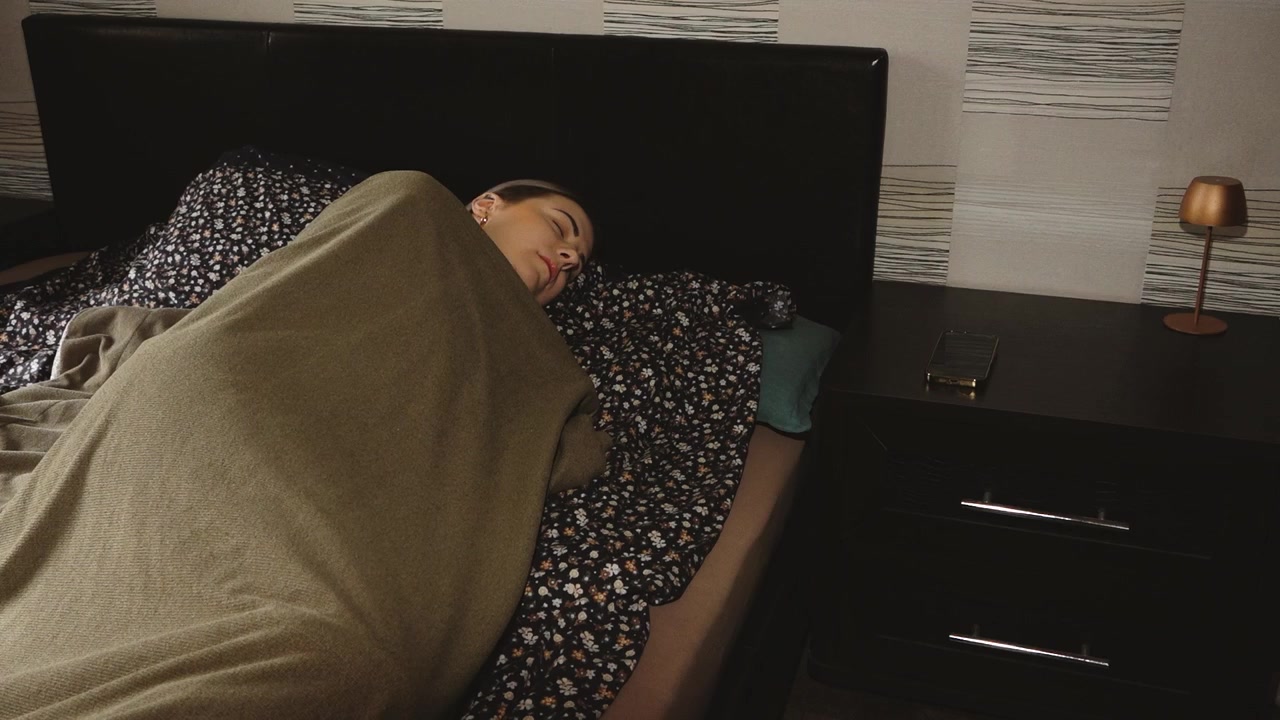 Žena leží pod dekou v posteli a natahuje se pro chytrý telefon na nočním stolku, čímž vytváří scénu klidného životního stylu v ložnici.