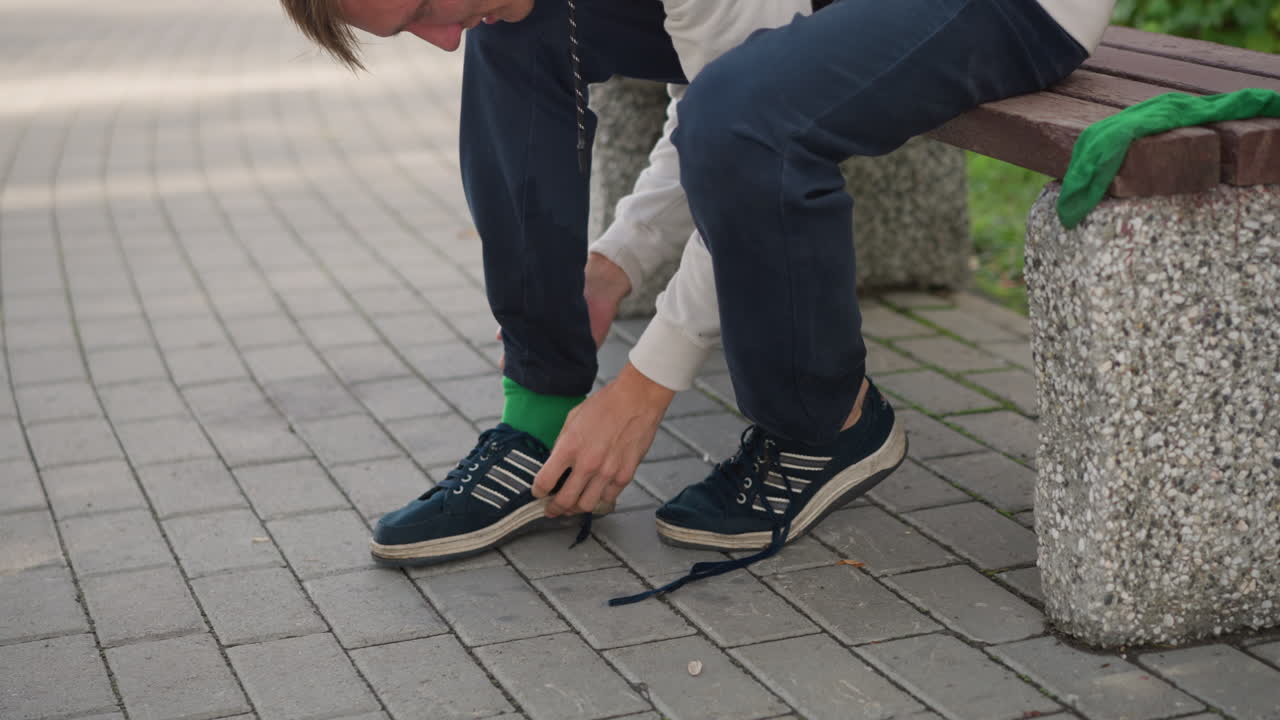 primer plano de una persona poniéndose un calcetín verde en un banco; un individuo se pone un calcetín en el pie descalzo tras quitarse la zapatilla; las manos ajustan la tela y los dedos; pavimento de adoquines y cielo nublado de fondo; recuperación tranquila