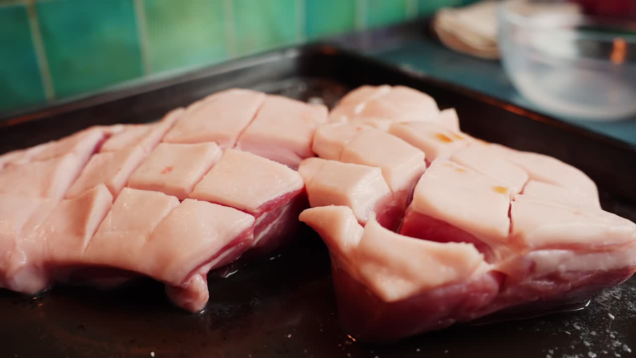 塩漬けされ、切り込みが入れられた豚バラ肉が暗いローストパンの上に置かれ、カリカリのクラックリングになるよう準備されている。家庭の台所での調理準備のクローズアップ、縦位置。