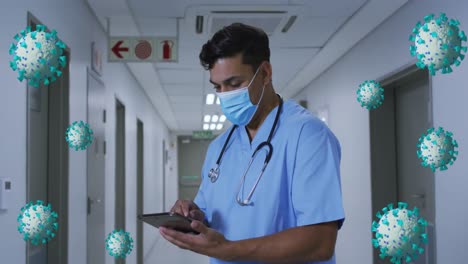 Animación-De-Células-Azules-De-Coronavirus-Moviéndose-Sobre-Un-Médico-Masculino-Con-Mascarilla-Usando-Una-Tableta-En-El-Hospital