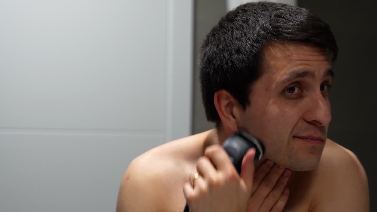 Man carefully shaving face with electric razor in bathroom mirror, focused expression