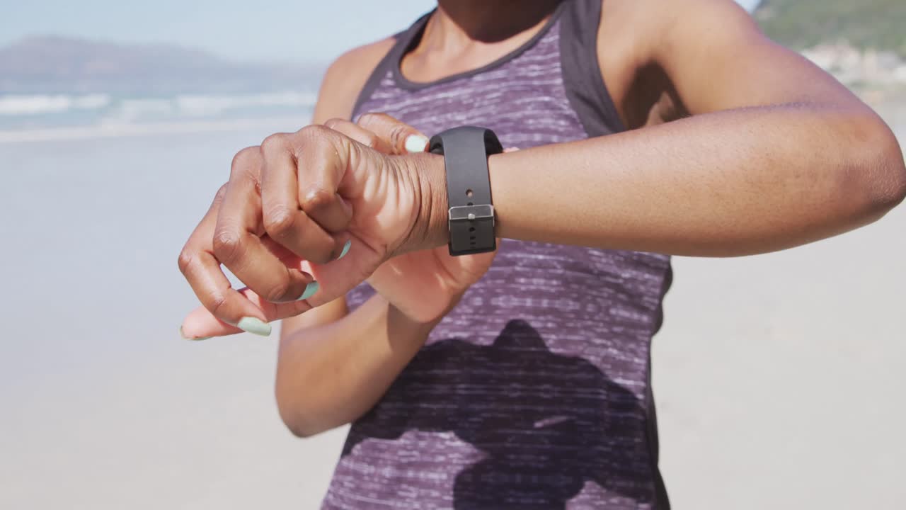 Kvinde af blandet etnicitet kigger på sit smartwatch på stranden med blå himmel i baggrunden.