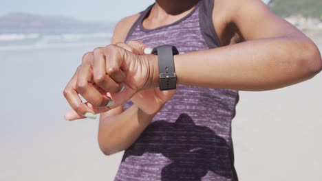 Mujer-De-Raza-Mixta-Mirando-Su-Reloj-Inteligente-En-La-Playa-Y-El-Fondo-Del-Cielo-Azul