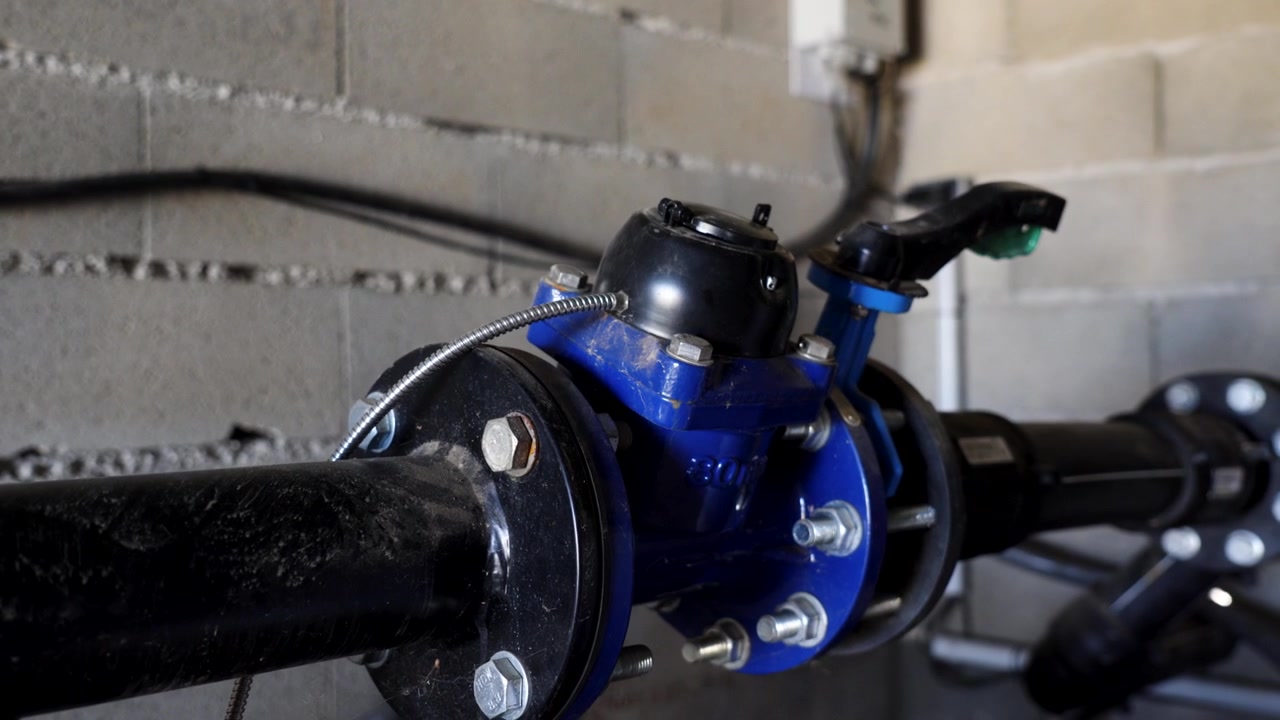 Primer plano de un contador de agua industrial azul de alta resistencia y una válvula de control instalados en una tubería con bridas en un sótano o cuarto de servicio
