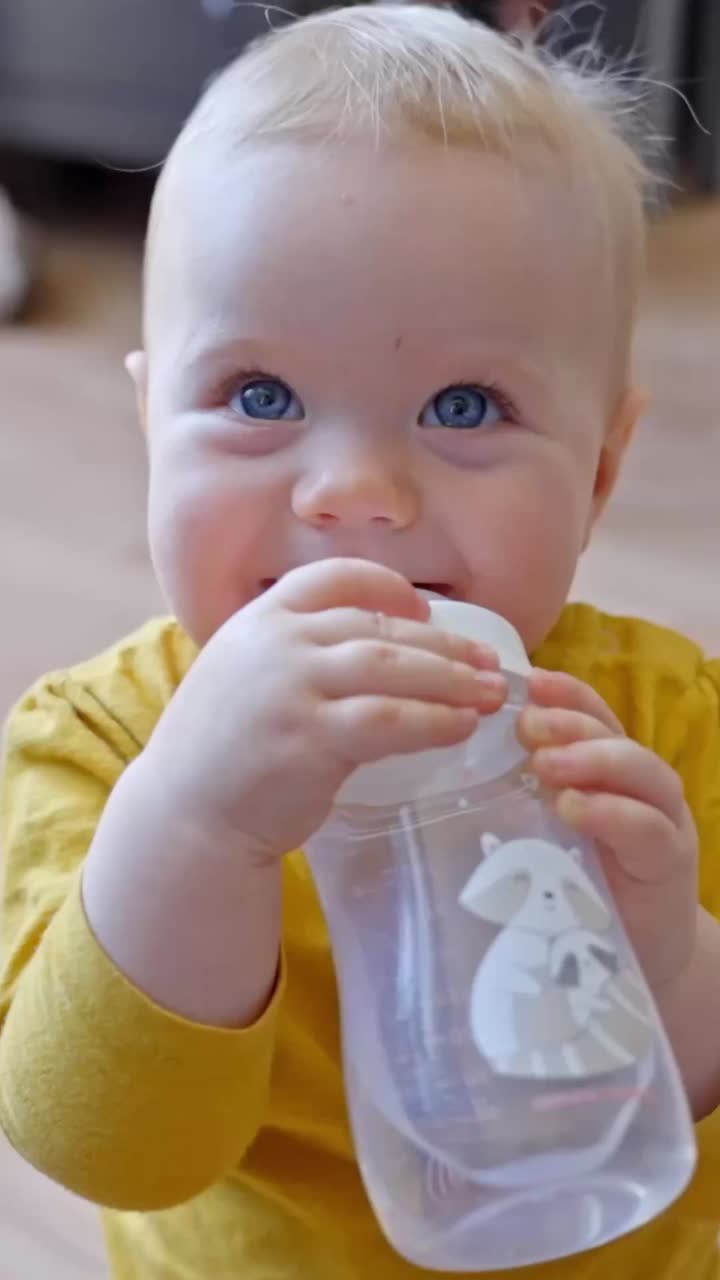 Baby holding water bottle and drinking while looking curiously indoors, vertical