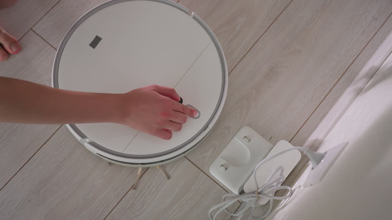 Overhead view of person operating robot vacuum on wooden floor, hand pressing control buttons while device charges near wall socket, blending household technology with everyday home cleaning