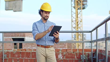 Arquitecto-Joven-Caucásico-De-Buen-Aspecto-Con-Casco-Parado-En-El-Techo-Del-Sitio-De-Construcción-Usando-Un-Dispositivo-De-Tableta,-Tocando-Y-Desplazándose.