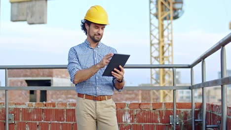 joven arquitecto caucásico de buen aspecto con casco de pie en el techo del sitio de construcción usando un dispositivo de tableta, tocando y desplazándose.