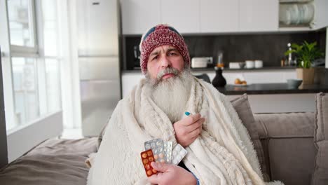 retrato de un anciano enfermo con una barba gris y exuberante en un sombrero rojo y un cuadrilátero blanco sosteniendo un termómetro y un conjunto de pastillas mientras está sentado en un sofá moderno en un apartamento